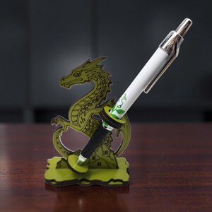 May include: A green dragon-shaped pen holder featuring a cutout design with black line details. A white and black pen with a green leaf graphic is inserted into the circular holder attached to the dragon figure, which stands on a flat base.