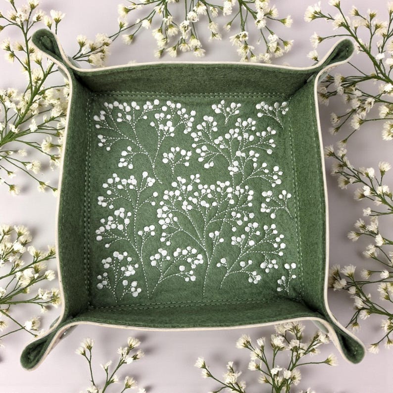 May include: A square, sage green felt tray with a white floral embroidery design. The tray has a cream-colored trim and curved handles. The tray is surrounded by white flowers.