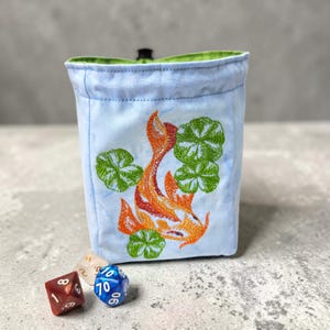 May include: A light blue dice bag with an embroidered orange koi fish and green lily pads. The bag has a green interior and a black drawstring closure. Three multi-sided dice are in front of the bag.