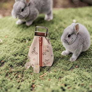 Puede incluir: Una pequeña bolsa beige con cremallera y asa metálica, adornada con un estampado floral, descansa sobre musgo verde. Dos conejos grises están en el fondo. Dados con el número uno están junto a la bolsa.