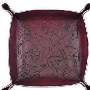 Puede incluir: Una bandeja de cuero burdeos con un diseño de hadas y flores. La bandeja tiene forma cuadrada con esquinas redondeadas y bordes curvos. El diseño de hadas y flores está cosido con hilo negro.