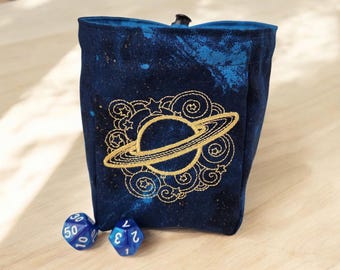 May include: A blue velvet dice bag with a gold embroidered design of a planet with rings. The bag is sitting on a wooden surface with two blue dice beside it.