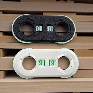 May include: Two oval-shaped counters, one black and one white, each with two circular openings and a series of green numbers. The black counter displays "0 0", while the white counter shows "9 19". The counters are set against a wooden background.