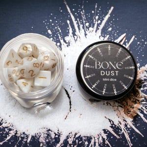 Puede incluir: Un pequeño frasco blanco con tapa negra que contiene dados blancos con números dorados. El frasco está etiquetado como "BONE DUST Mini dice".
