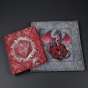 May include: A red, embossed leather-bound book with a silver D20 die design, next to a gray felt dice tray. The tray features an embroidered pink dragon perched on a red D20 die. A unique set for tabletop gaming.
