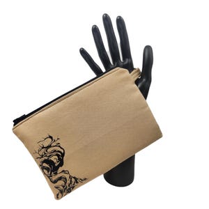 May include: A tan canvas pouch with a black zipper and a black abstract tree design on the front. The pouch is held by a black mannequin hand against a white background. The pouch is rectangular.
