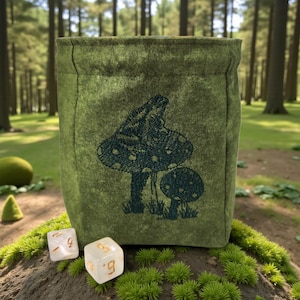 May include: A green fabric dice bag with a dark green embroidered mushroom design. Two dice with gold numbers are in front of the bag. The bag is sitting on a bed of green moss.