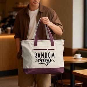 May include: Cream-colored tote bag with a burgundy base, trim, and handles. The bag features a front pocket with a zipper and the words "RANDOM crap" in black script. The bag is held by a person.