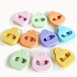 May include: A collection of ten colorful, rubbery, earplug-like objects. Each is a different pastel color, including pink, green, orange, purple, and yellow. Each has a letter and number printed on it.