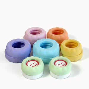May include: A collection of seven circular silicone bottle caps in pastel colours including purple, pink, orange, yellow, blue, and two light green caps featuring a red smiling face graphic on the top surface. Each cap has a scalloped edge.
