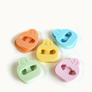 May include: Five colorful, rubbery, oval-shaped objects in orange, yellow, green, blue, and pink. Each has a rectangular opening in the center and a letter and number printed on the top. The objects are arranged on a white background.