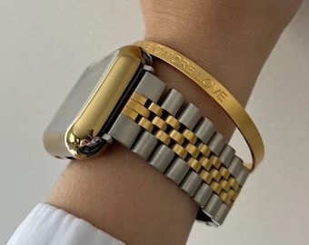 Puede incluir: Un reloj dorado y plateado con una correa a juego se lleva en la muñeca. También se lleva una pulsera dorada con la inscripción "MORE LOVE". El reloj y la pulsera destacan sobre un fondo blanco.