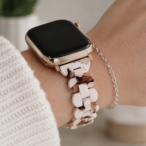 May include: A smartwatch with a metallic gold-tone case and a black screen, featuring a link-style watch band made of cream and brown tortoiseshell-patterned resin. The watch is worn on a wrist alongside a delicate, thin silver-tone chain bracelet.
