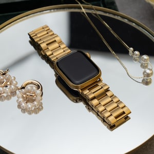 Può includere: Uno smartwatch color oro con cinturino in metallo abbinato è appoggiato su una superficie a specchio, accompagnato da orecchini di perle e una delicata collana. Un portagioie verde e fiori bianchi sono sullo sfondo. L'orologio ha uno schermo nero.