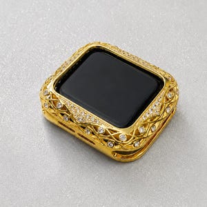 May include: A gold-toned smartwatch protective case featuring an intricate lattice design and embedded clear crystals surrounding a black screen. The metallic frame has a polished finish with a detailed, repeating pattern that covers the edges.