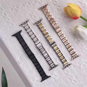 Könnte beinhalten: Vier Metall-Uhrenarmbänder in Schwarz, Silber, Gold und Roségold. Jedes Armband besteht aus rechteckigen Gliedern und ist für eine Smartwatch konzipiert. Die Armbänder werden auf einer weißen, strukturierten Oberfläche mit einer gelben und weißen Blume im Hintergrund präsentiert.