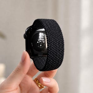 Może przedstawiać: Czarny smartwatch z plecionym, elastycznym paskiem o teksturowanym splocie. Urządzenie posiada okrągły czujnik na tylnej obudowie, a pasek wykonany jest z ciągłego, elastycznego i rozciągliwego materiału o jednolitej czarnej kolorystyce.