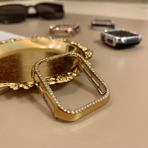 Può includere: Una custodia dorata per Apple Watch con una lunetta bordata di piccoli cristalli trasparenti. La custodia è appoggiata su un vassoio dorato e ornato. Altri accessori per orologi e occhiali da sole sono sullo sfondo.