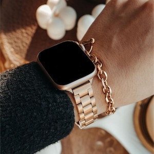 Puede incluir: Un reloj inteligente dorado con pantalla negra se lleva en la muñeca. El reloj tiene una correa dorada a juego y se combina con una pulsera de cadena dorada. El reloj y la pulsera se colocan sobre un fondo oscuro.