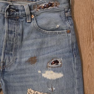 May include: Light blue denim jeans with decorative embellishments. Features include a leopard print patch, sequin detailing on the waistband, and distressed patches. The jeans have a classic five-pocket design.