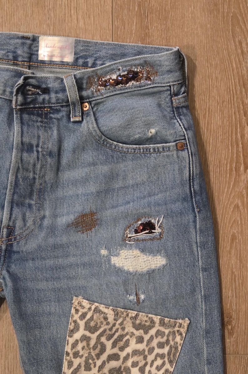May include: Light blue denim jeans with decorative embellishments. Features include a leopard print patch, sequin detailing on the waistband, and distressed patches. The jeans have a classic five-pocket design.