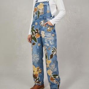 May include: Light blue denim dungarees with floral patches and distressed details. The dungarees feature a bib pocket, adjustable straps, and brown leather boots. The floral patches include yellow, brown, and green colours.