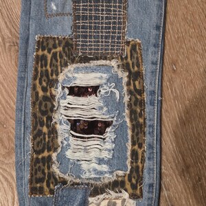 May include: Close-up of distressed blue denim jeans with multiple patches. Patches include leopard print, a woven fabric, and a sequined section. The jeans have ripped sections and frayed edges, showcasing a unique, patched design.
