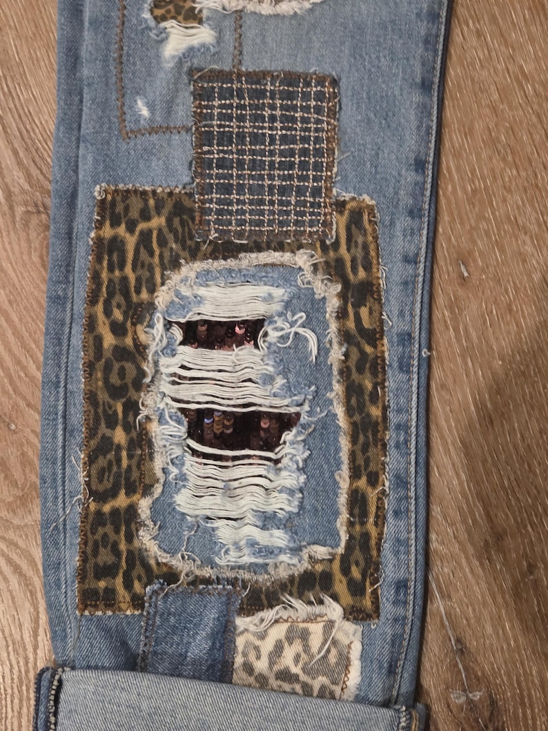 May include: Close-up of distressed blue denim jeans with multiple patches. Patches include leopard print, a woven fabric, and a sequined section. The jeans have ripped sections and frayed edges, showcasing a unique, patched design.