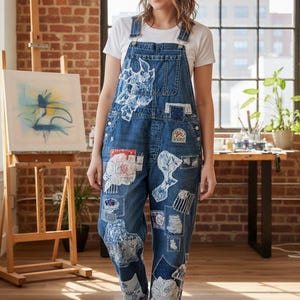 May include: Blue denim dungarees with white patches and floral designs. The dungarees have adjustable straps and a bib pocket. The person is wearing a white t-shirt and white trainers. The background includes a brick wall, a window, and an easel with a painting.