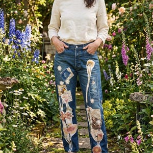 Könnte beinhalten: Blaue Jeans mit floralen und Bienen-Patchwork-Designs. Die Jeans hat einen geraden Bein-Schnitt und ist mit Stoffflicken in Rosa, Beige und Braun verziert. Die Person trägt ein cremefarbenes Langarmshirt.