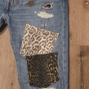 May include: Light blue denim jeans with leopard print pocket patches and distressed details. The jeans feature ripped areas with visible repairs and patches, showcasing a unique, customized design.