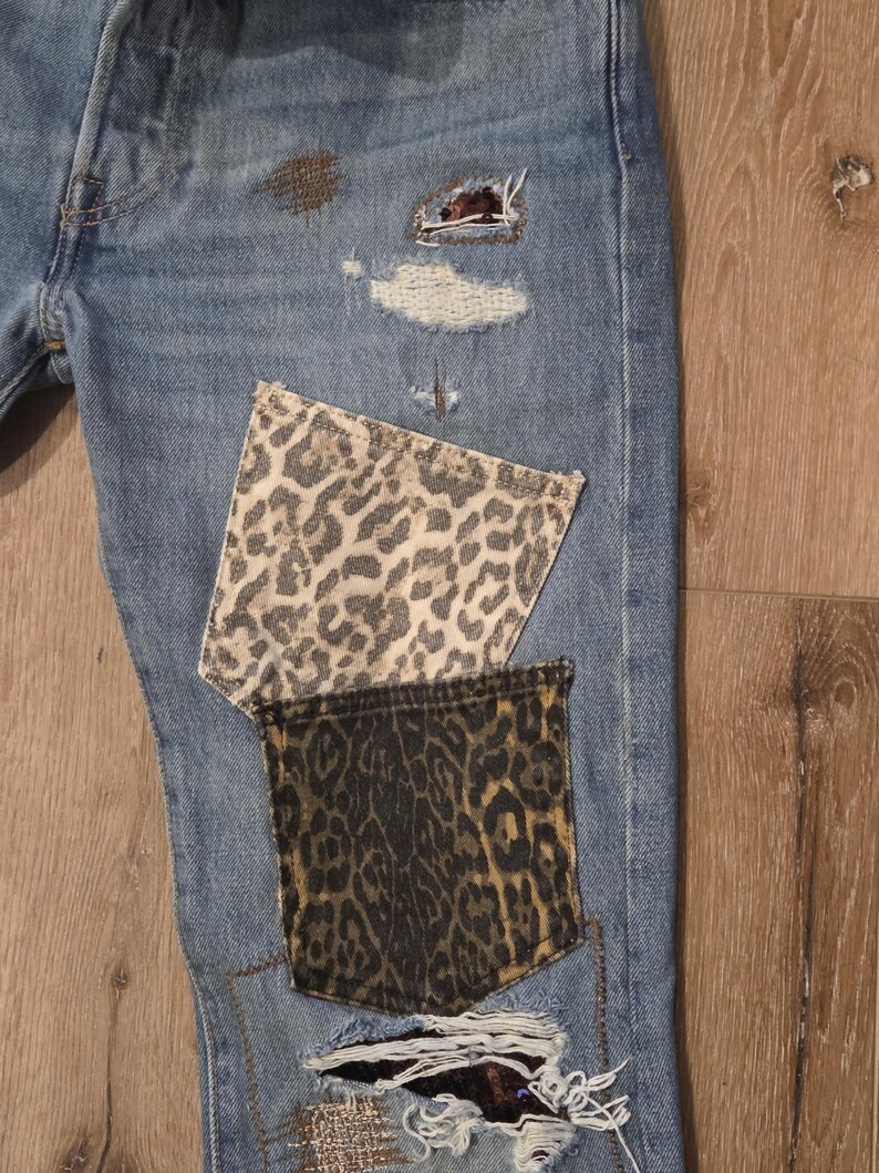 May include: Light blue denim jeans with leopard print pocket patches and distressed details. The jeans feature ripped areas with visible repairs and patches, showcasing a unique, customized design.