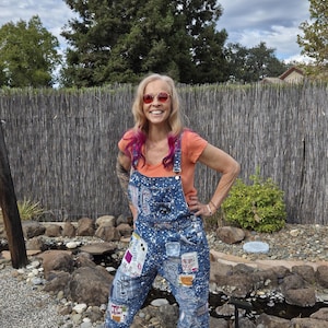 May include: Blue denim overalls featuring a white paint splatter pattern, various fabric patches, and distressed details. The overalls have a bib front with metal hardware, side buttons, and multiple pockets. An orange short-sleeve t-shirt is worn underneath.