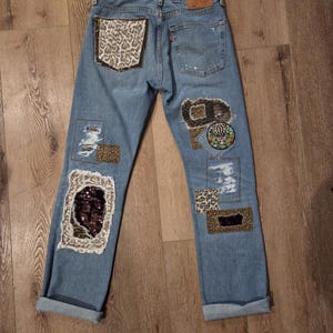 May include: Light blue denim jeans featuring various patches and embellishments. Patches include leopard print, sequins, and embroidered designs. The jeans have a distressed appearance with rolled-up cuffs and a classic five-pocket design.