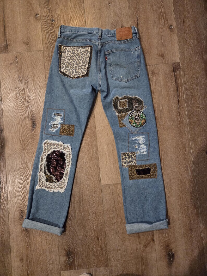 May include: Light blue denim jeans featuring various patches and embellishments. Patches include leopard print, sequins, and embroidered designs. The jeans have a distressed appearance with rolled-up cuffs and a classic five-pocket design.