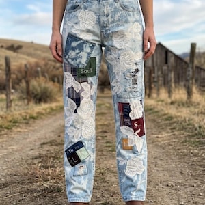 May include: Light blue denim jeans with white floral lace appliques and patches. The jeans have distressed details and patches with the words "Soul" and "Yo Joy". Brown leather boots complete the look.