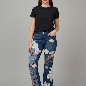 Op de afbeelding: Een vrouw draagt een zwart t-shirt met ronde hals en een blauwe spijkerbroek. De jeans heeft decoratieve patches met bloemen- en hartmotieven en witte kanten details. Ze draagt witte sneakers.
