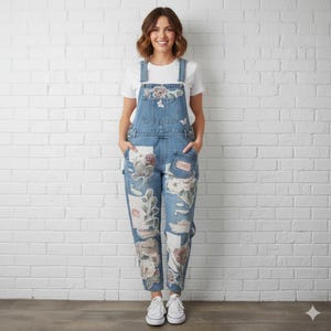 May include: Light blue denim dungarees with floral patches and a white t-shirt. The dungarees have a bib pocket, front pockets, and adjustable straps. The floral patches are in pink, white, and green. The model is wearing white trainers.