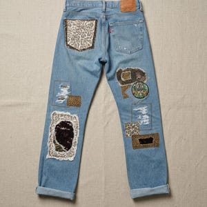 May include: Light blue denim jeans featuring various patches. Patches include leopard print, sequined fabric, and embroidered designs. The jeans have a distressed appearance with ripped details and rolled-up hems.