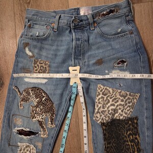 May include: Light blue denim jeans with leopard print patches and a leopard applique. The jeans have distressed details and a measuring tape laid across the front. The waist measures approximately 25 cm.