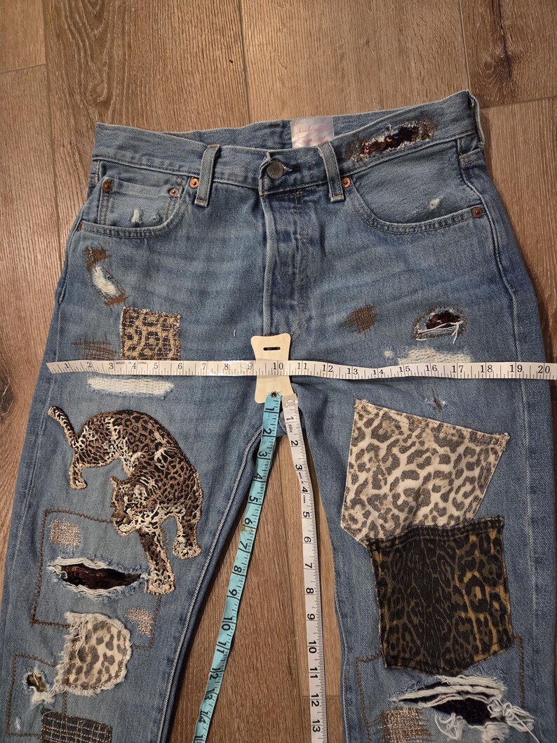May include: Light blue denim jeans with leopard print patches and a leopard applique. The jeans have distressed details and a measuring tape laid across the front. The waist measures approximately 25 cm.