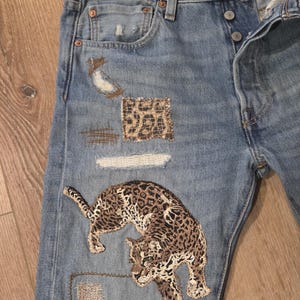May include: Light blue denim jeans with distressed details and patches. Features a large embroidered leopard design in brown and tan. Additional patches include leopard print and distressed fabric.