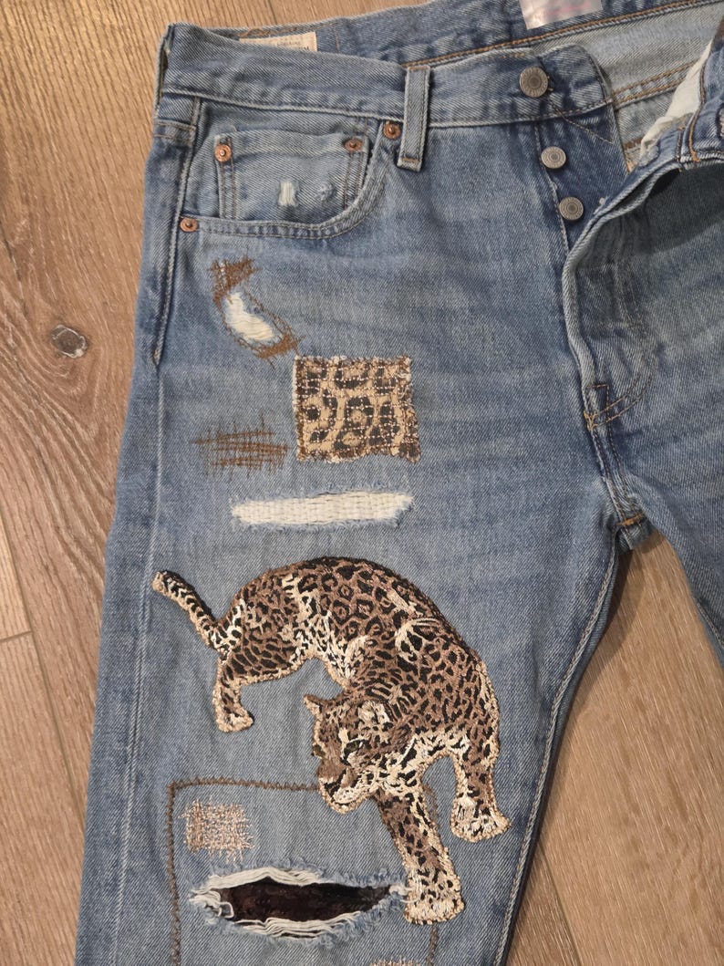 May include: Light blue denim jeans with distressed details and patches. Features a large embroidered leopard design in brown and tan. Additional patches include leopard print and distressed fabric.