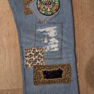May include: Light blue denim jeans with decorative patches. Patches include leopard print, a circular embroidered patch with a tiger, and a distressed area. The jeans have a unique, patched design.