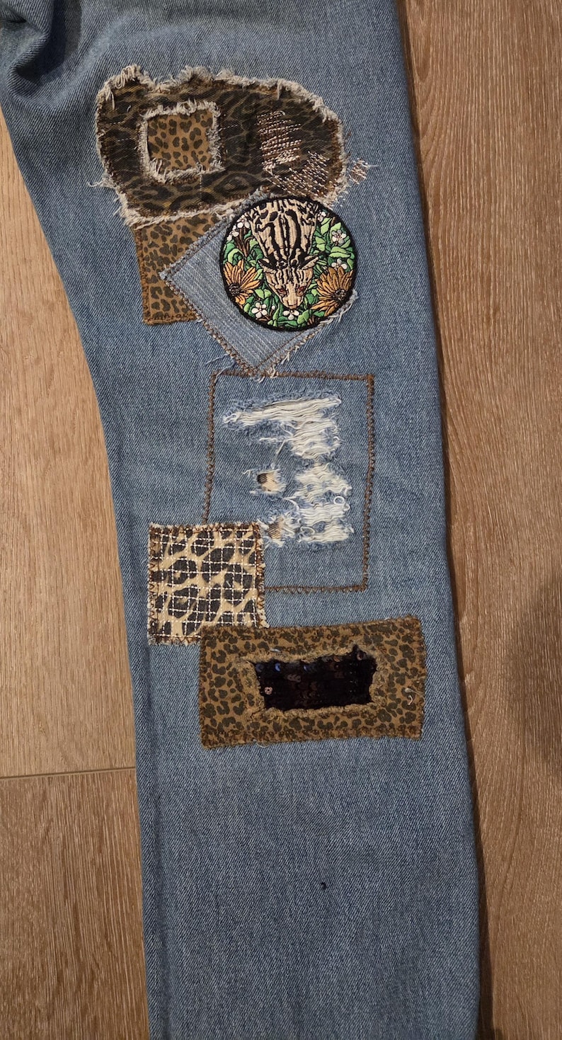 May include: Light blue denim jeans with decorative patches. Patches include leopard print, a circular embroidered patch with a tiger, and a distressed area. The jeans have a unique, patched design.
