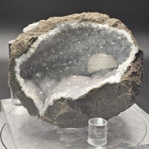 May include: A geode with a grey exterior and a white crystal interior. The geode is open, revealing the crystals inside. The geode is sitting on a clear display stand.