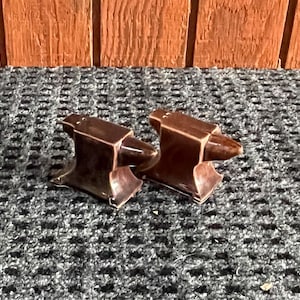 May include: Two brown ceramic anvil figurines. Each figurine has a rectangular base, a central block, and a pointed horn. The anvils are displayed on a textured gray surface, with a wooden background.