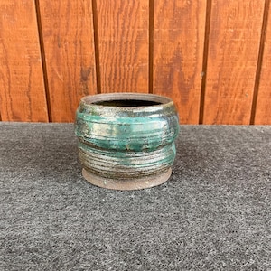 May include: A small, round, ceramic planter with a dark brown glaze and green stripes. The planter is sitting on a gray surface.