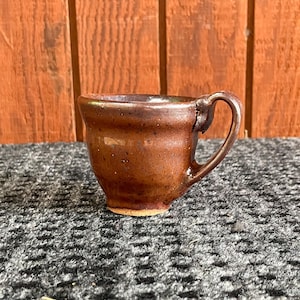 May include: A small, handmade brown ceramic mug with a handle. The mug has a rounded body and a slightly flared rim. The exterior is a mottled brown color with darker speckles. The mug sits on a textured surface.