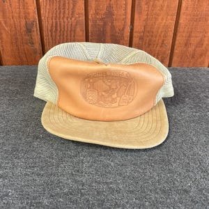 May include: Tan and beige trucker hat with a leather patch. The patch features an engraved design with the words "FINES TOOLS" and a graphic of a person and tools. The hat has a mesh back and a curved brim.